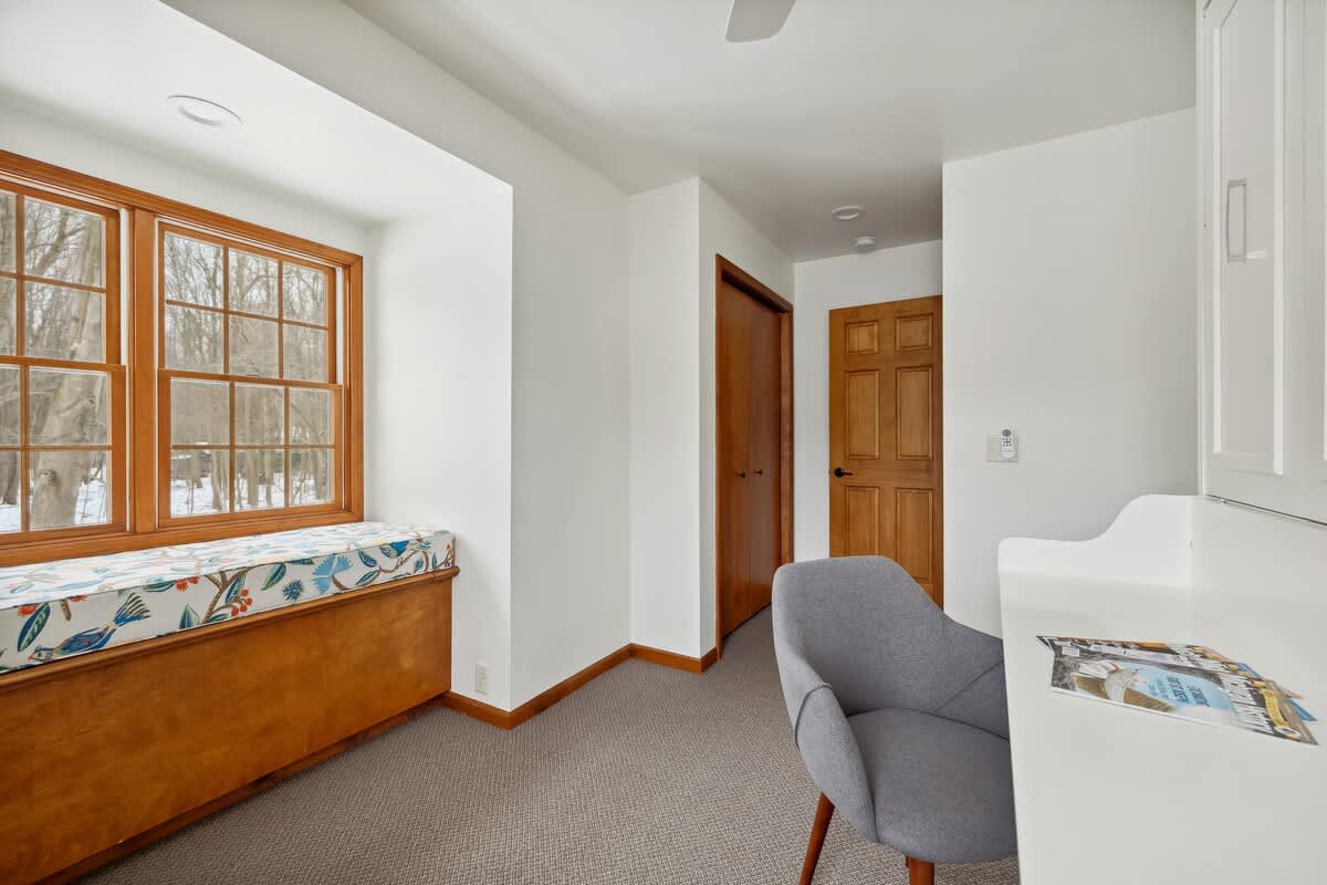A bright office space with a built-in window seat featuring a colorful patterned cushion, overlooking a snowy outdoor scene. A gray office chair and part of a white built-in desk are visible, with wooden trim throughout.