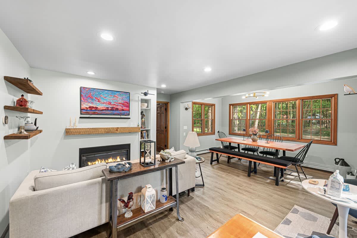 An open-concept living and dining area features a light-colored sectional sofa facing a fireplace and TV, with a rustic console table behind it. A long wooden dining table with benches and chairs is positioned by multiple wood-framed windows.