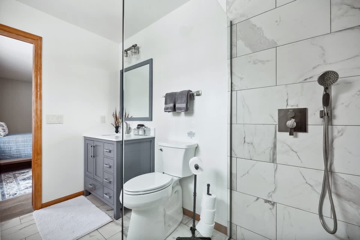View of bathroom-1 showing the toilet next to the grey vanity and the large walk-in shower with its glass panel. White marble-look tiles cover the shower walls and floor, with a small white rug in front of the vanity.