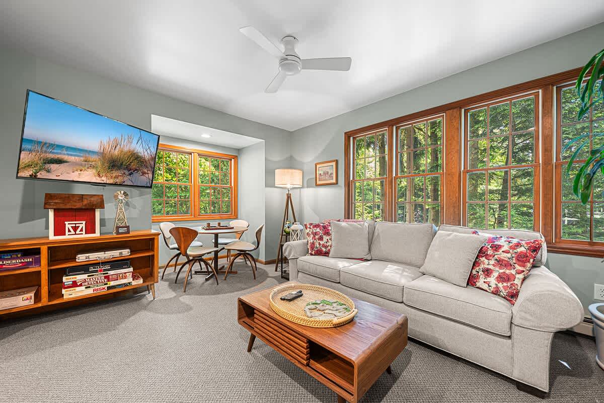 Cozy game room with light grey walls, carpeted flooring, and large windows offering views of the outdoors. Features a comfortable sofa, a wall-mounted TV above a media console with board games, and a small dining or game table with chairs.