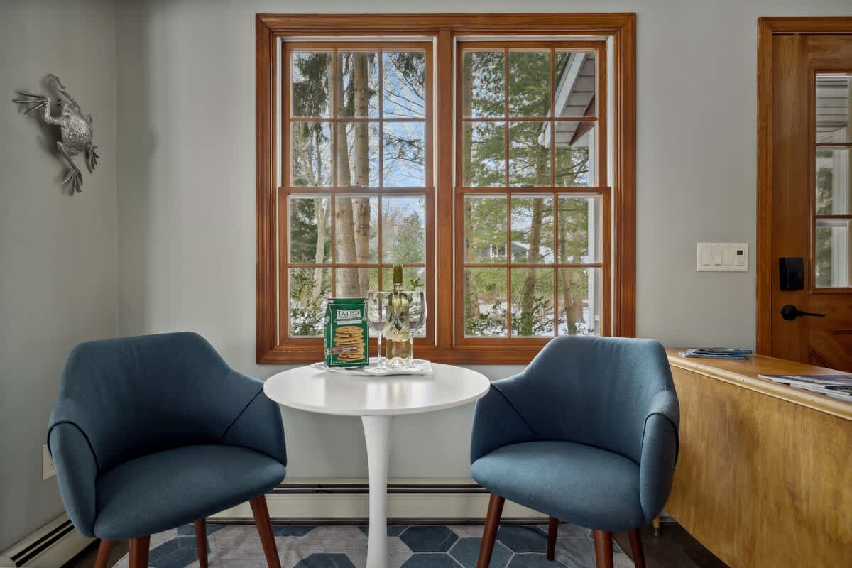 A close-up of a bright dining nook shows a white round table with two blue upholstered chairs on a blue and white geometric rug, framed by large wood-trimmed windows overlooking a snowy outdoor scene.