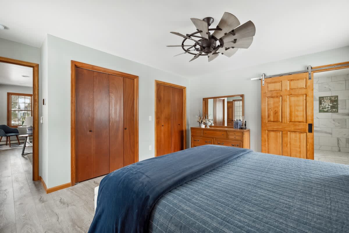 View of bedroom-1 highlighting two wooden closet doors and a large wooden dresser with a mirror. A wooden barn door slides open to reveal an en-suite bathroom, all set against light blue walls and wood-look flooring.