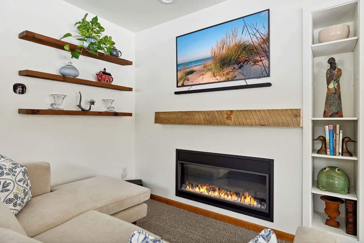 A cozy living room features a modern electric fireplace with a rustic wood mantel, topped by a wall-mounted TV displaying a beach scene. Floating wooden shelves with decorative items adorn the adjacent wall, next to a built-in white bookshelf.