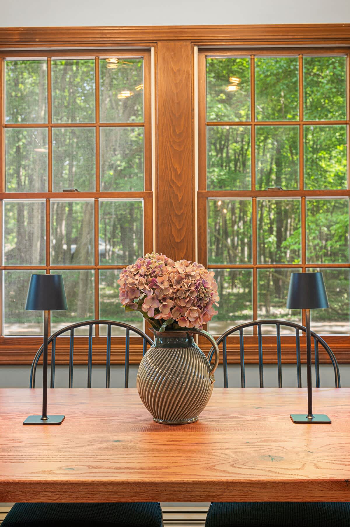 A rustic wooden dining table is adorned with a decorative vase of flowers and two modern black table lamps, set against a backdrop of large wood-framed windows offering a lush green outdoor view.