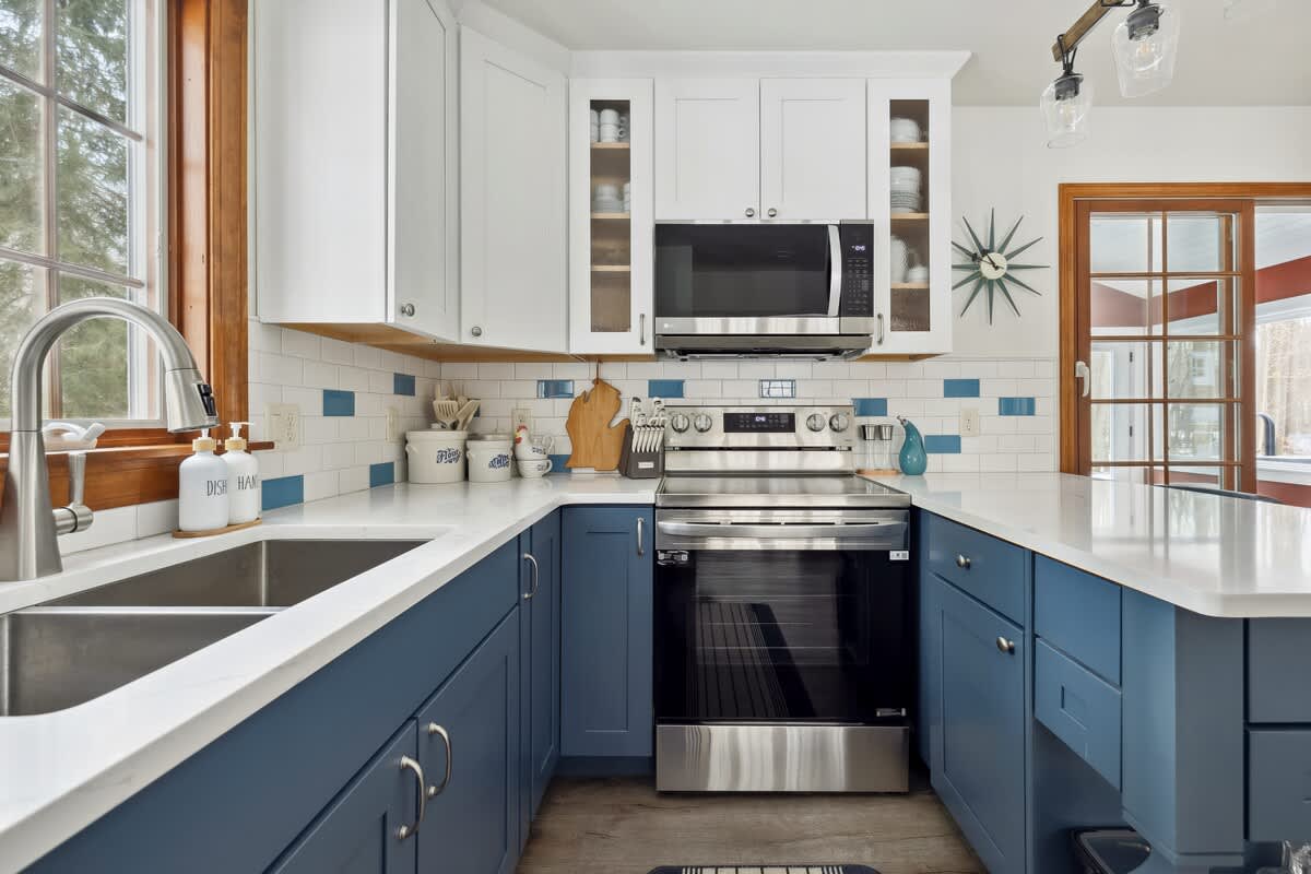A well-lit kitchen corner features a double-basin stainless steel sink under a wood-framed window, and a stainless steel range with a microwave above. Blue and white shaker cabinets and a subway tile backsplash with blue accents complete the area.