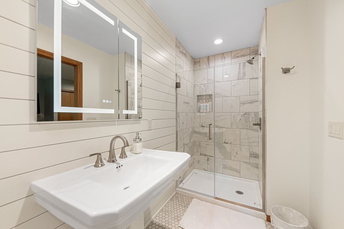 A modern bathroom features a white pedestal sink with a brushed nickel faucet, an illuminated mirrored medicine cabinet, and shiplap-style white walls. A glass-enclosed shower with white marble-patterned tiles is visible to the right.