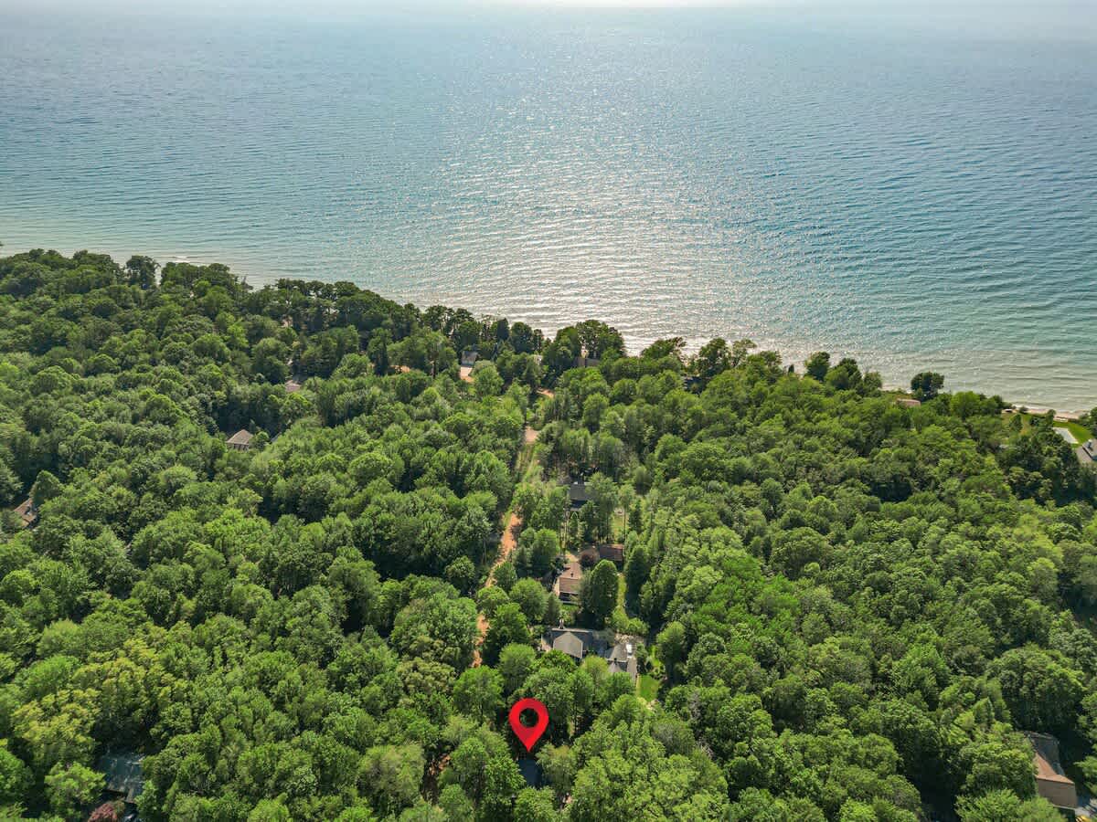 An aerial view captures the property nestled deep within a dense, vibrant green forest, with a vast expanse of blue water, likely a lake or ocean, visible in the background. A red location pin marks the house's position amidst the lush tree canopy.