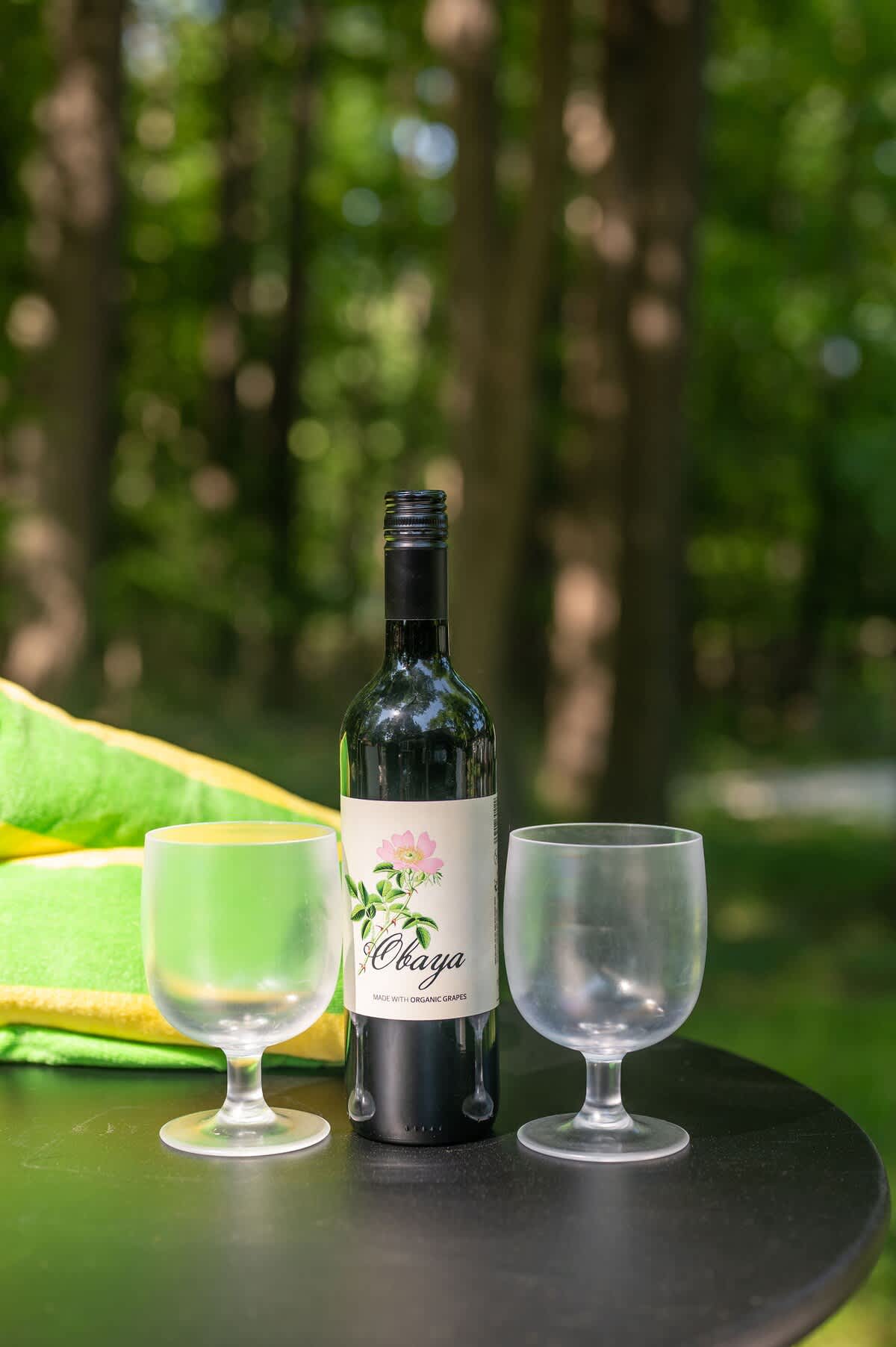 A close-up of a bottle of "Obaya" organic wine and two glasses on a black outdoor table, set against a lush green forest background. A bright green towel is partially visible.