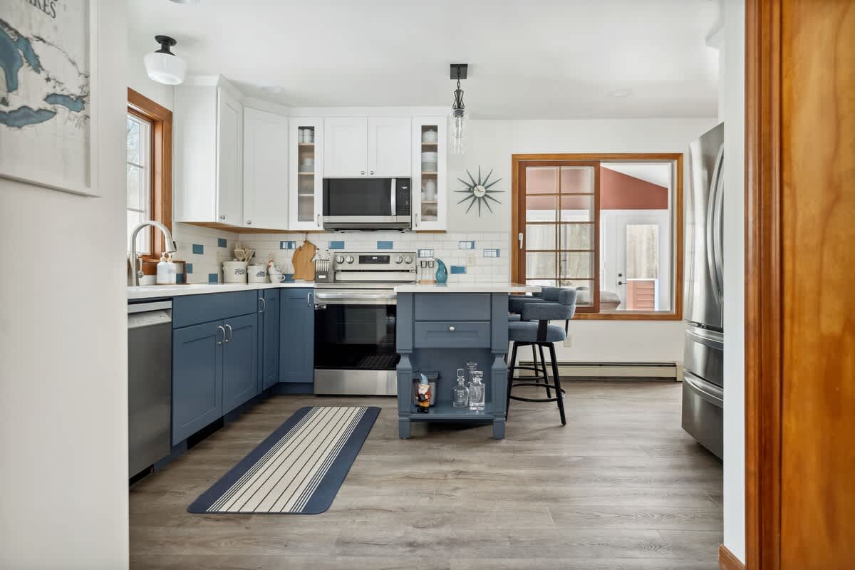 A modern kitchen features two-toned blue and white shaker-style cabinets, white quartz countertops, and a subway tile backsplash with blue accents. A central island with seating and stainless steel appliances complete the space.