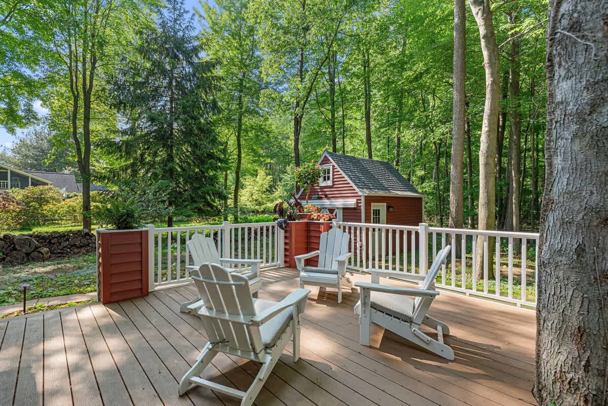 A spacious wooden deck furnished with several white Adirondack chairs and a white railing, offering a serene view of a lush green forest. A small red shed is nestled among the trees in the background.