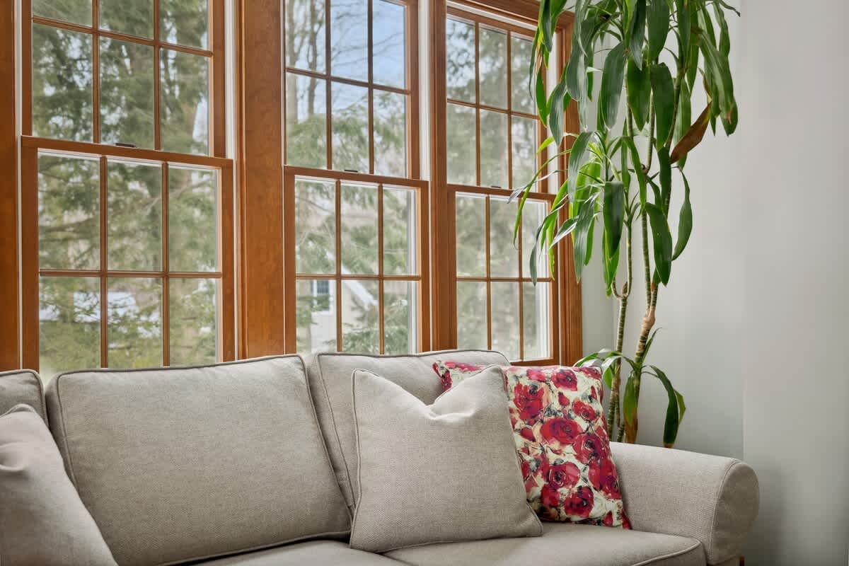 A close-up of the comfortable light gray sofa in the living room, adorned with light-colored and red floral throw pillows. Large wooden-framed windows provide a bright view of the surrounding trees.