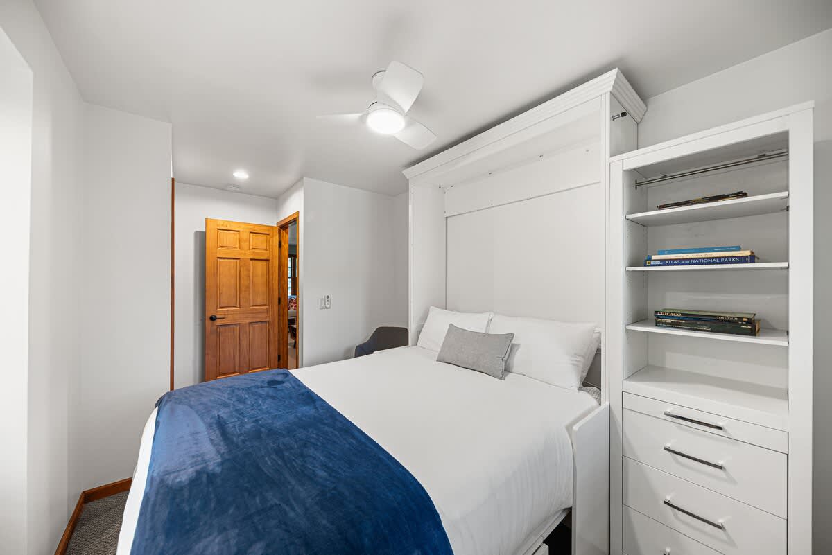 A compact bedroom featuring a white Murphy bed with a blue throw blanket, flanked by white built-in shelving and drawers. A wooden door is visible in the background, and a ceiling fan is overhead.