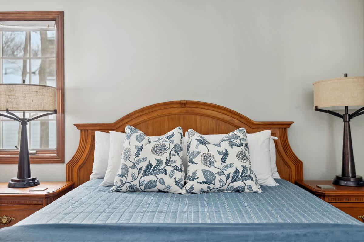 Close-up view of the bed in bedroom-1, showcasing a classic wooden headboard, light blue quilted bedding, and two decorative pillows with a blue and white floral pattern.