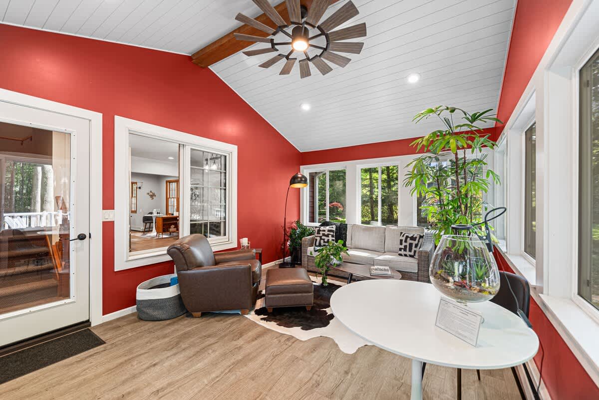 Bright sunroom with vibrant red walls, white vaulted ceiling with a large ceiling fan, and light wood-look flooring. Features a cozy seating area with a leather armchair, sofa, and a white round table, all surrounded by large windows offering outdoor views.