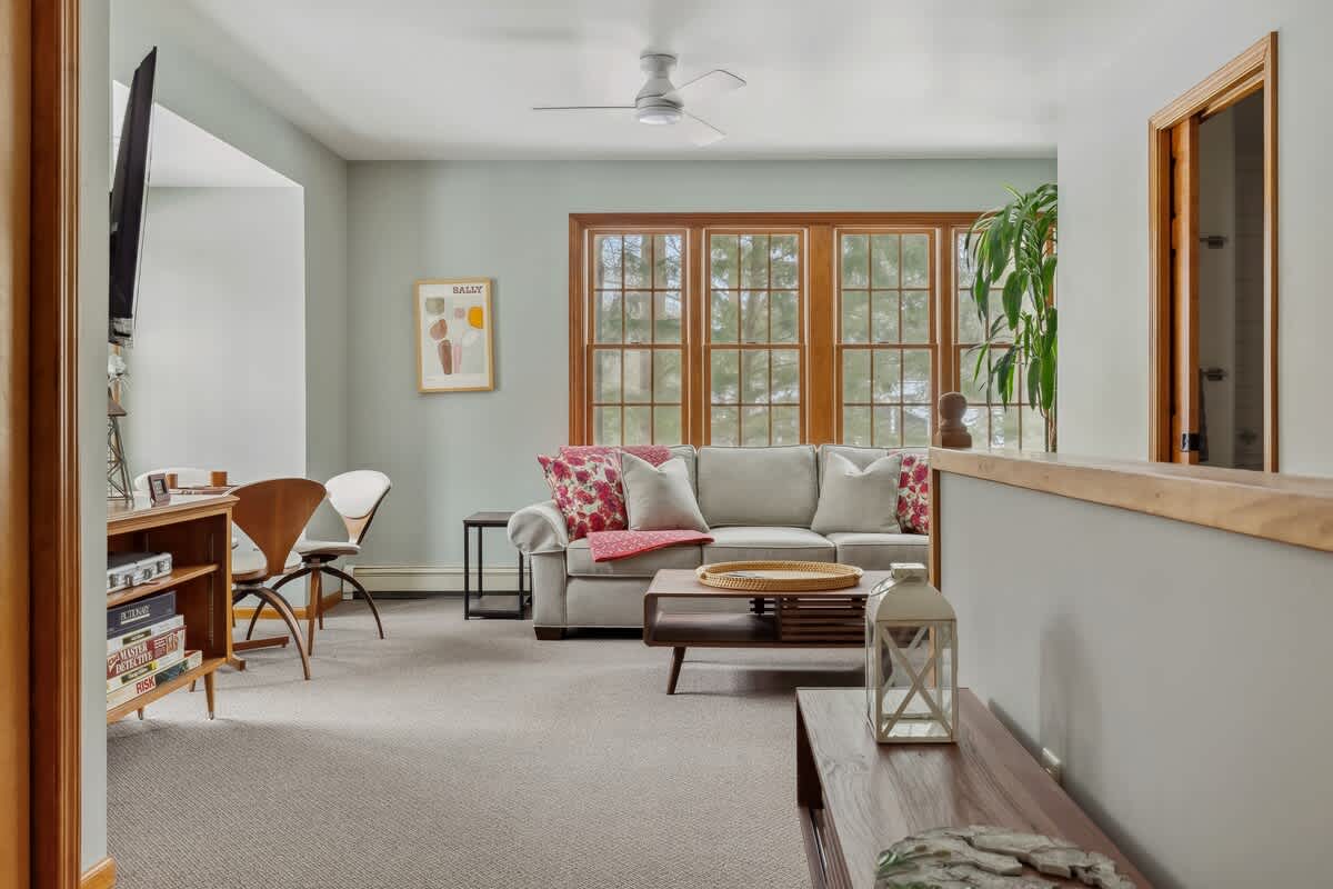 A spacious living room with light gray walls and carpet features a comfortable light gray sofa adorned with red floral pillows and a throw. A wooden coffee table sits in front of the sofa, and large wooden-framed windows offer a view of the outdoors.