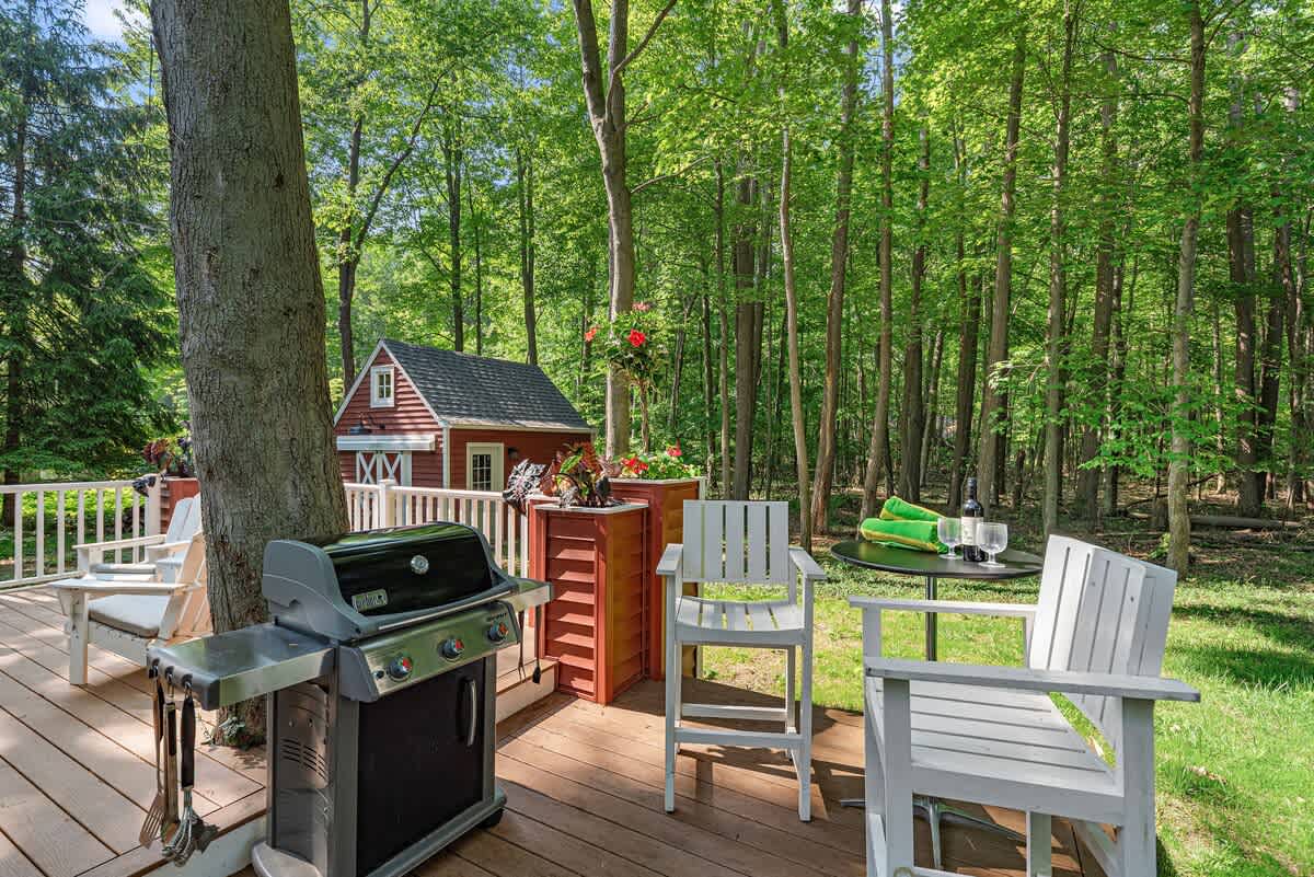 A wooden outdoor deck featuring a gas grill, white Adirondack-style chairs, and a small black table with wine glasses, all surrounded by a dense green forest. A red shed is visible in the background.