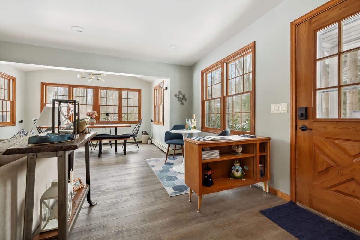 This bright and open-concept living space features light gray walls and wood-look flooring, creating an airy and welcoming atmosphere. It includes a wooden console table and a small round dining table, with multiple large windows framed by natural wood trim offering picturesque views of the surrounding greenery.
