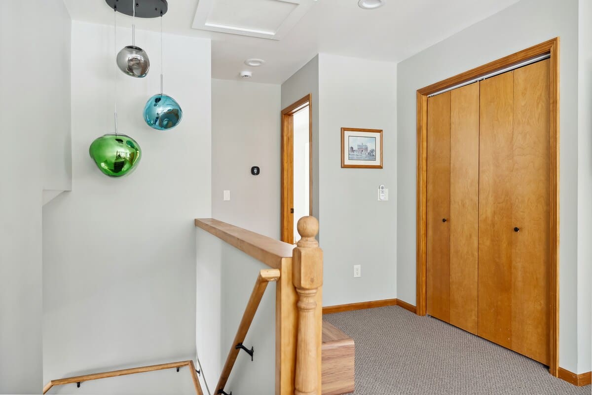 Upstairs landing area with light grey walls, carpeted flooring, and a wooden staircase railing. Features unique multi-colored glass pendant lights hanging from the ceiling and wooden closet doors.