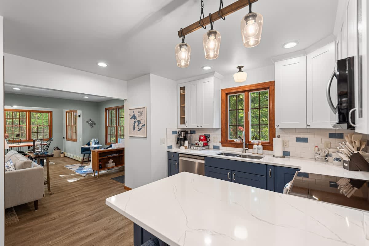 An open-concept kitchen with blue and white cabinetry, white countertops, and a subway tile backsplash flows into a bright living and dining area. A large window above the sink offers an outdoor view.
