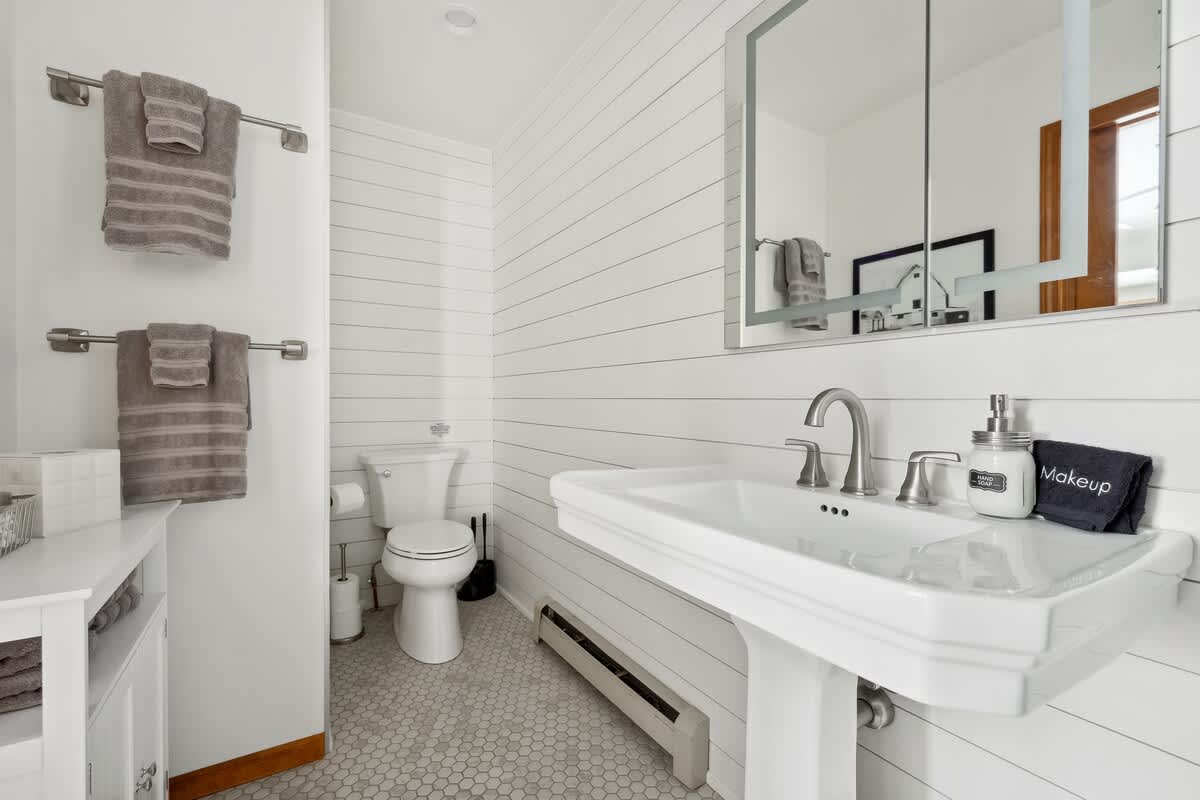 A bright bathroom features white shiplap walls, a white pedestal sink with a brushed nickel faucet, and a toilet. Gray towels are neatly hung on a rack, and the floor is covered in small white hexagon tiles.