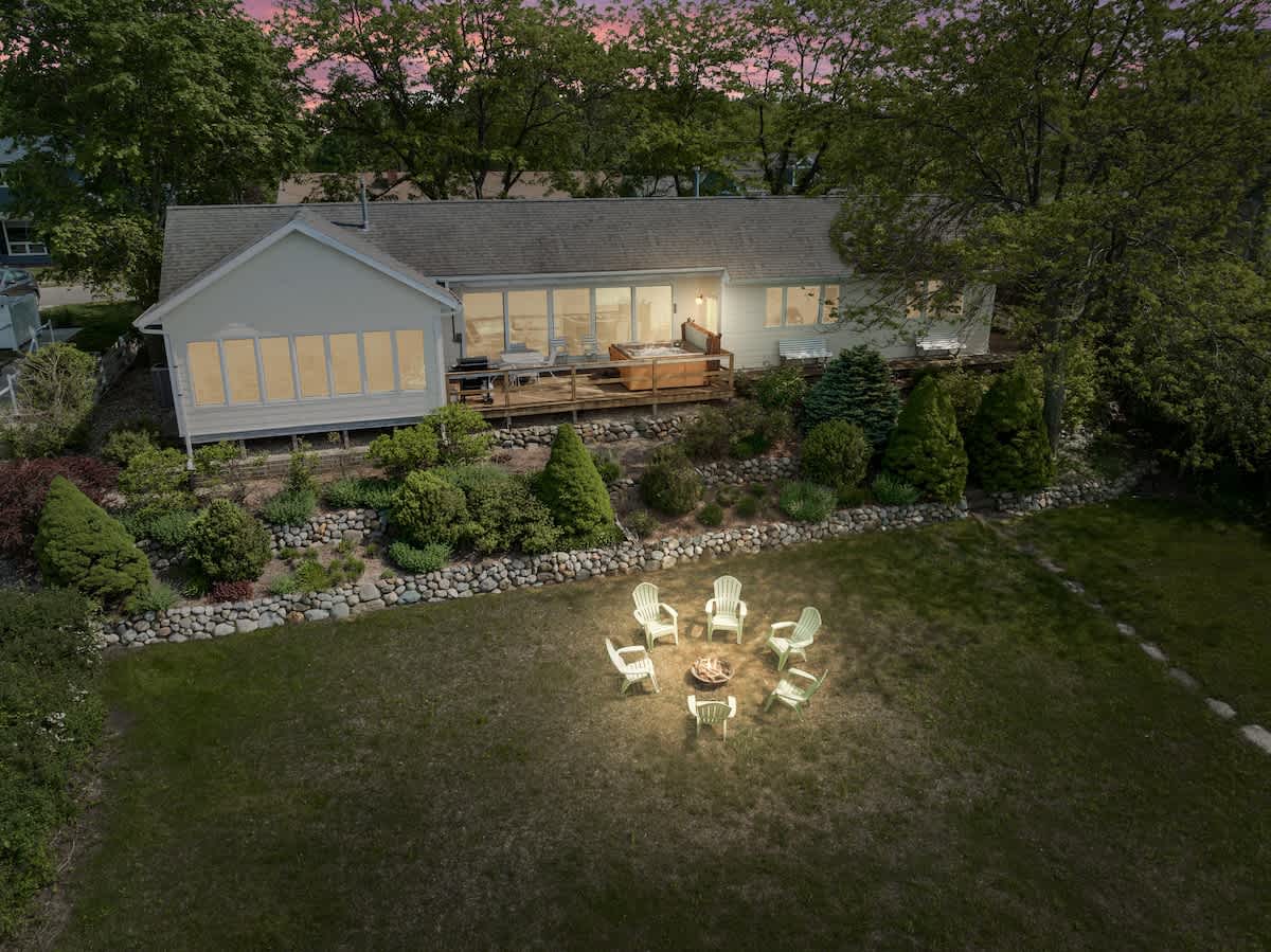 An aerial twilight view showcases the back of the house with a spacious deck, a hot tub, and a sunroom featuring large windows. Below, a glowing fire pit surrounded by Adirondack chairs illuminates the grassy yard, with the dark lake beyond.