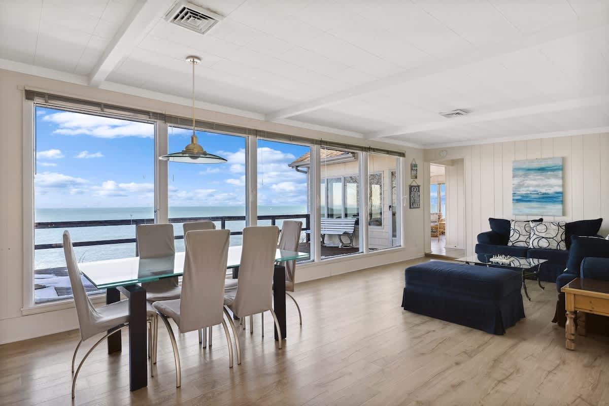 Open-concept living and dining area featuring light wood floors, a glass-top dining table with modern chairs, blue sofas, and large windows offering panoramic lake views.