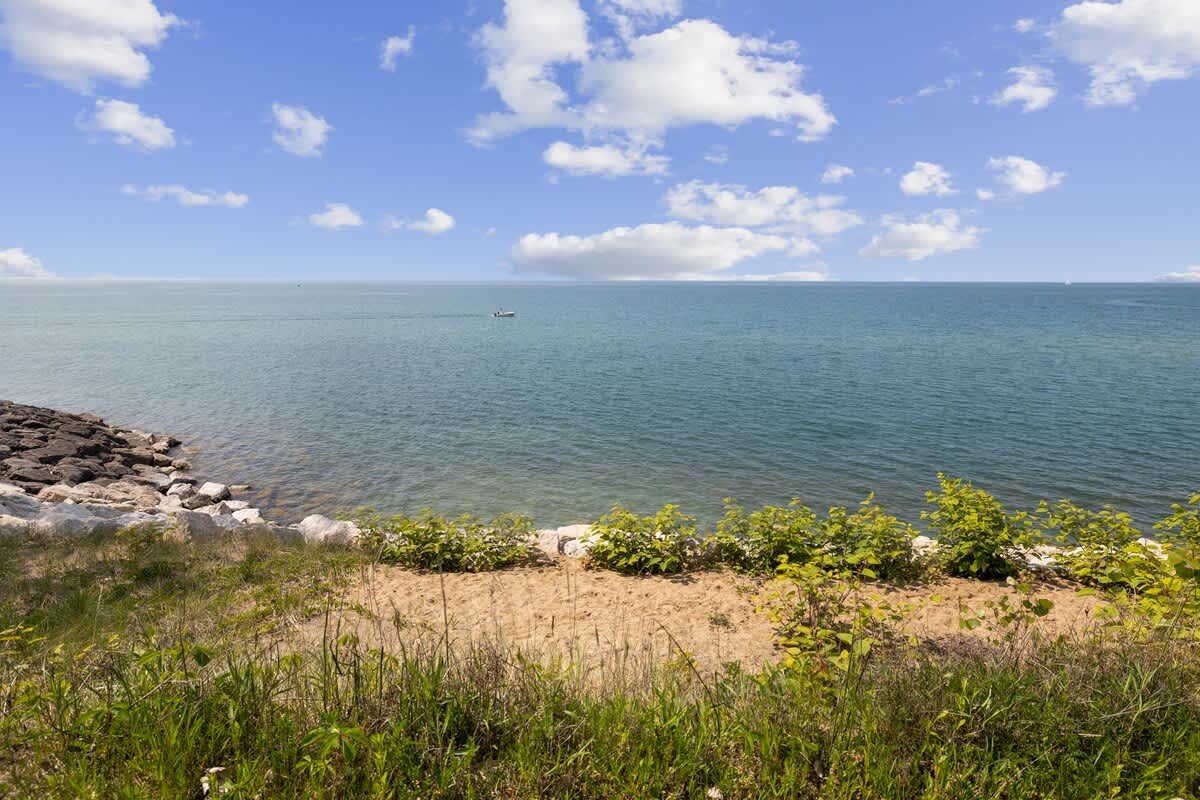A serene ground-level view captures the calm blue lake, with a small boat visible in the distance under a partly cloudy sky. The foreground features a natural sandy bank with sparse green vegetation, leading to a rocky shoreline.