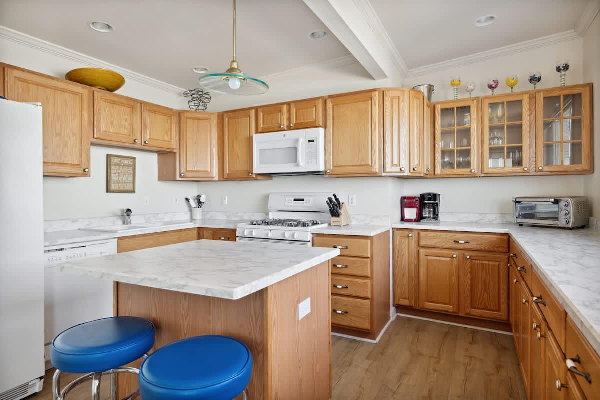 A spacious kitchen boasts light wood cabinets, white marble-look countertops, white appliances including a stove and dishwasher, a central island with blue stools, and a view towards a large window.