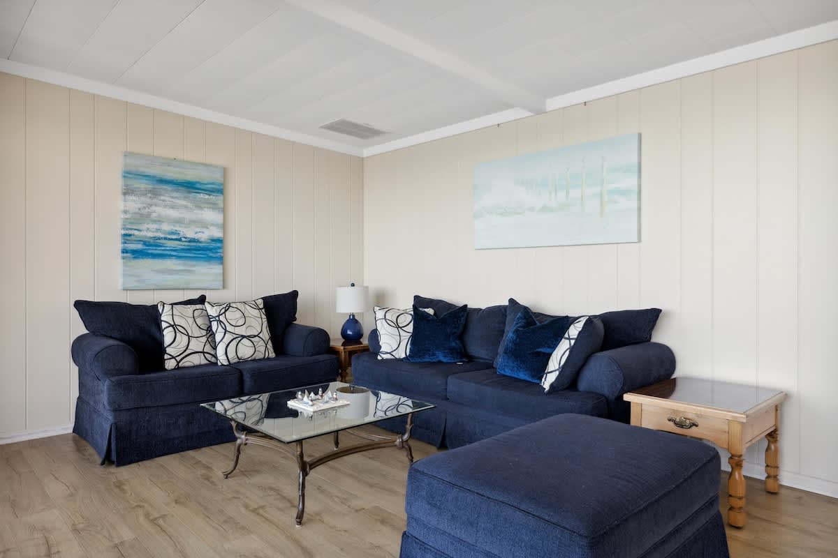 A cozy living room features two navy blue sofas adorned with patterned and solid blue pillows, a glass coffee table, light wood-look flooring, and two abstract blue and white artworks on the wall.