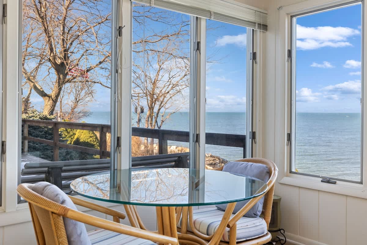 A bright sunroom dining nook features a round glass-top table and two wicker chairs, surrounded by large windows that offer an expansive, unobstructed view of the lake and the outdoor deck.