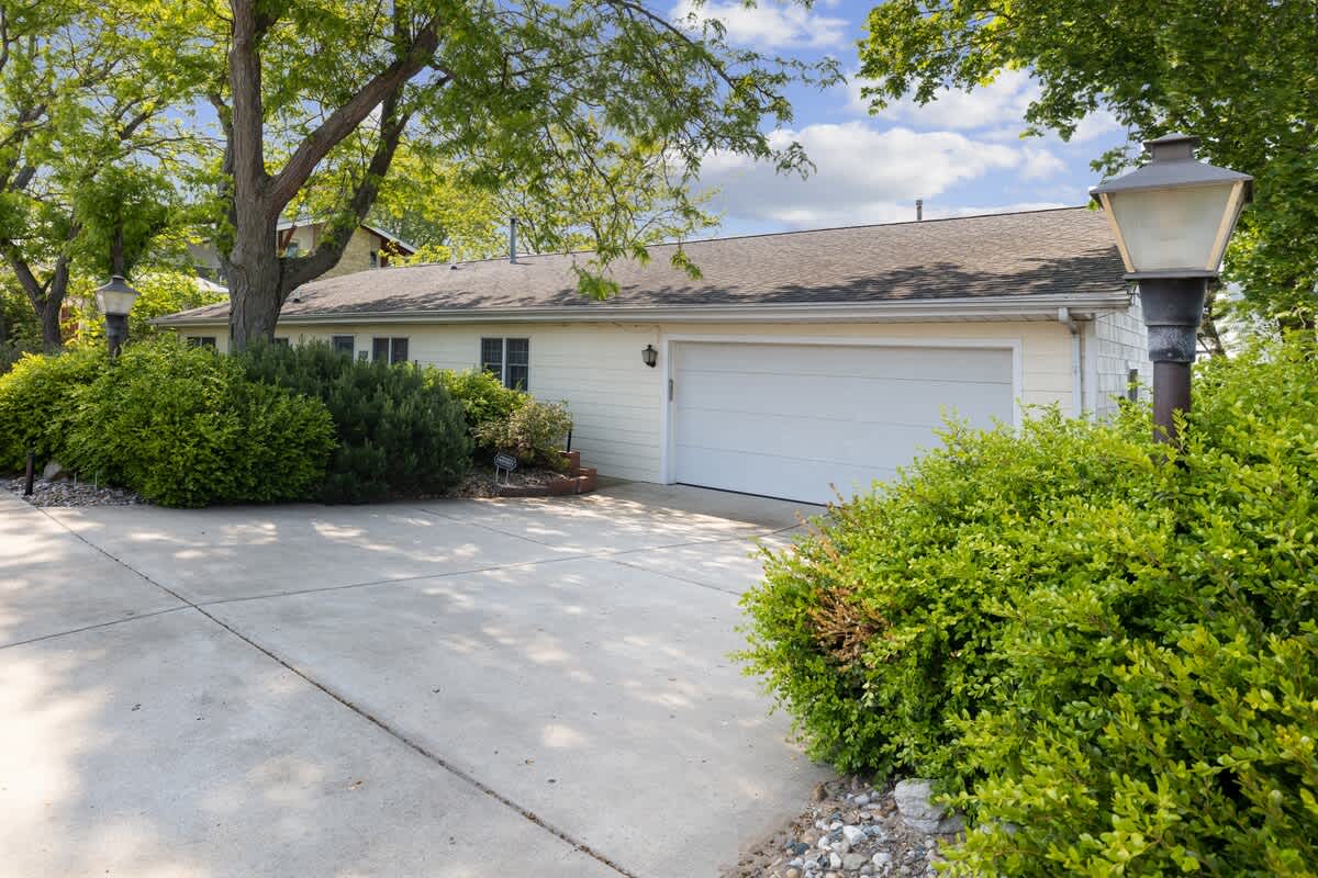 The front exterior of a single-story home features light-colored siding, a white two-car garage door, and a concrete driveway. Lush green bushes and mature trees frame the house, with a classic street lamp adding a touch of charm.