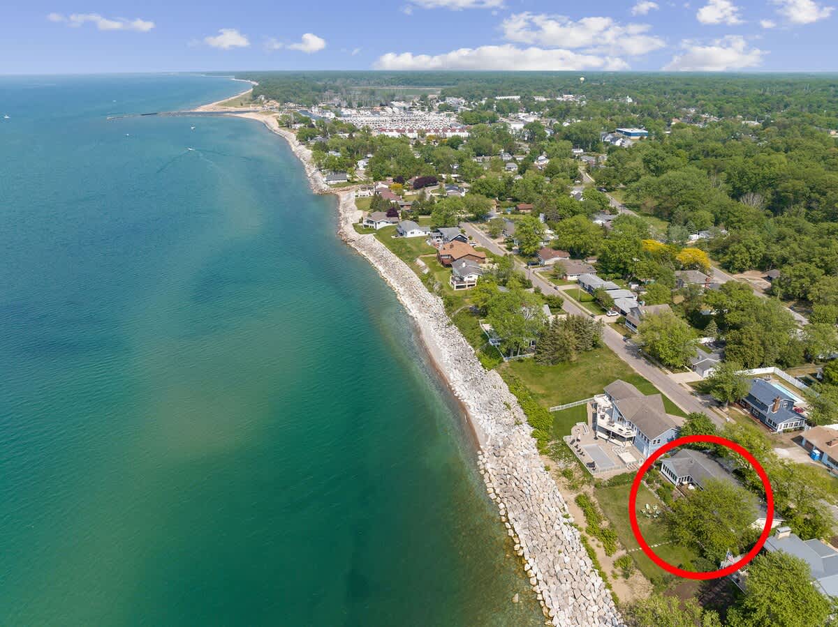 A high aerial shot captures the expansive coastline, featuring a vibrant blue lake and a residential town. The property, circled in red, is prominently situated beachfront, adjacent to a protective rocky breakwater.