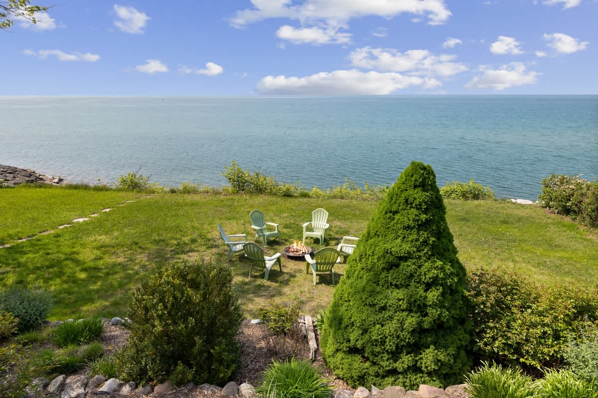A vibrant green lawn slopes down to a serene lake, featuring a circular fire pit area with six light-colored Adirondack chairs. The expansive blue water meets a clear sky, creating a picturesque outdoor gathering space.