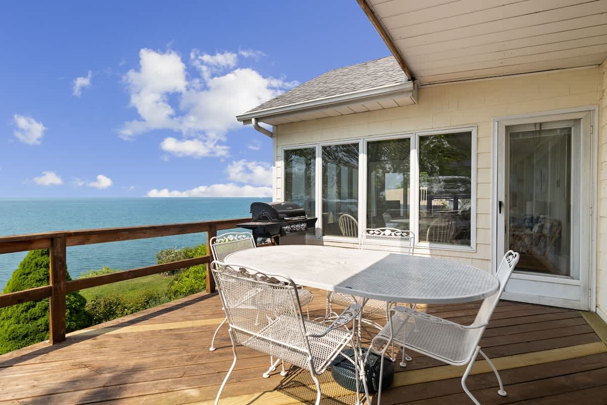 Spacious wooden deck furnished with a white metal dining set and a BBQ grill, offering an unobstructed, breathtaking view of the vast blue lake.
