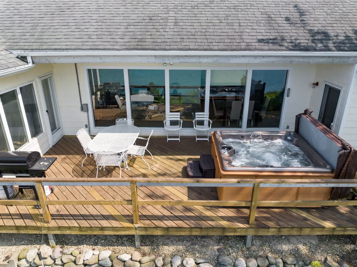 Overhead view of the expansive wooden deck featuring an outdoor dining area, a bubbling hot tub, and large sliding glass doors leading into the house, all overlooking the lake.