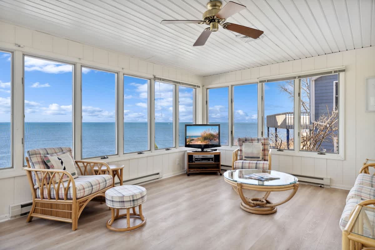Cozy sunroom with light wood floors, wicker armchairs, a glass coffee table, and a television, surrounded by large windows offering stunning lake views.