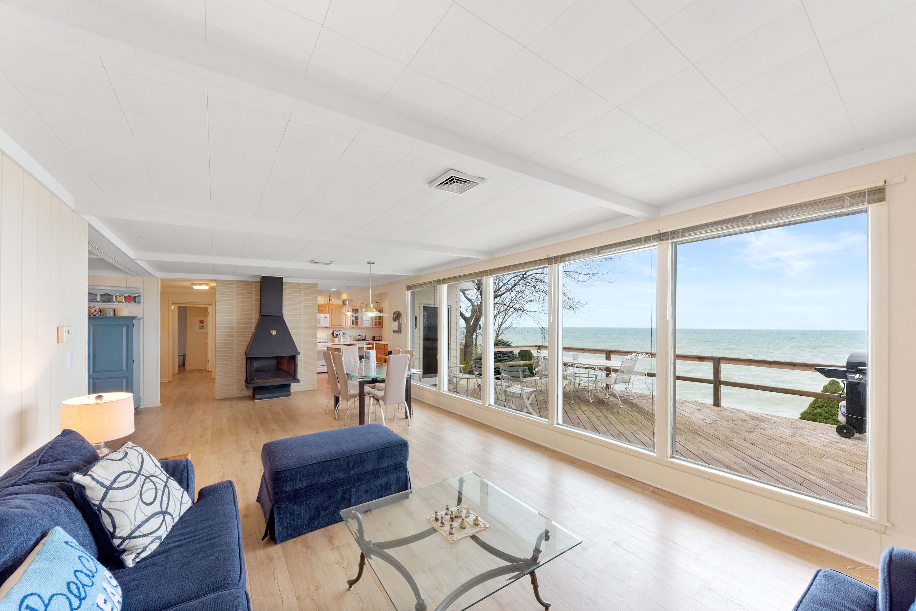 Spacious open-concept living and dining area with light wood floors, a prominent black fireplace, blue sofas, and a dining table, all with views of the lake through large windows.