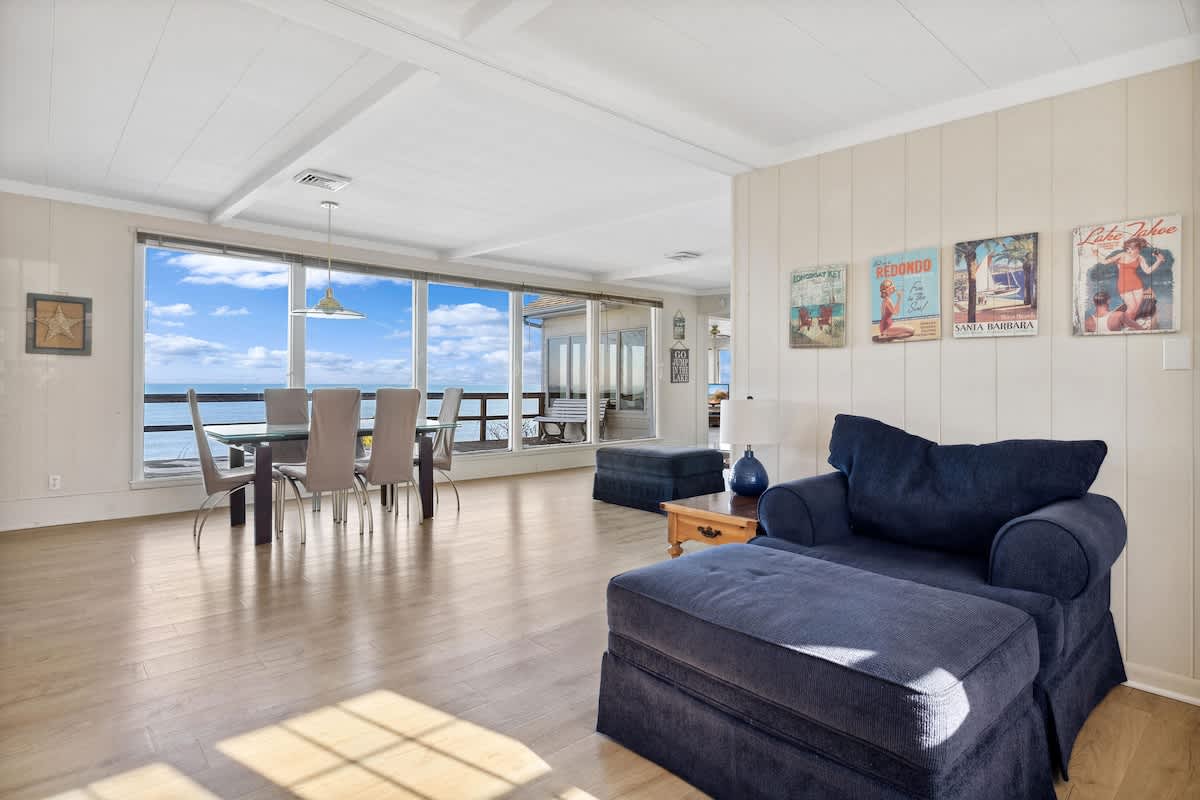 Expansive open-concept living and dining space with light wood floors, a blue sectional sofa, and a glass dining table, all illuminated by natural light from large windows overlooking the lake.