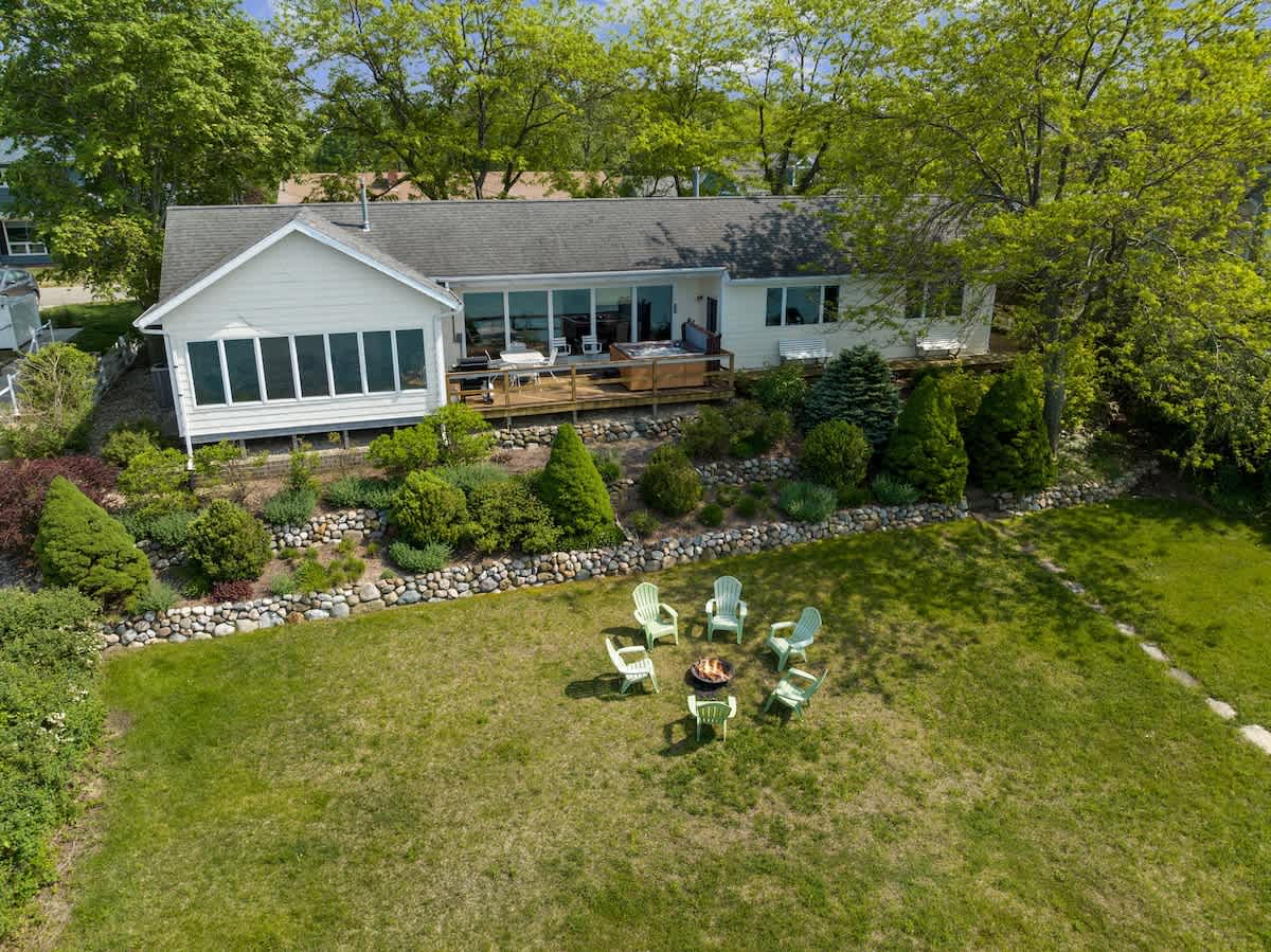 Aerial view of the back of the lakefront house, featuring a large green lawn with a fire pit surrounded by Adirondack chairs, and terraced landscaping leading to the lake.