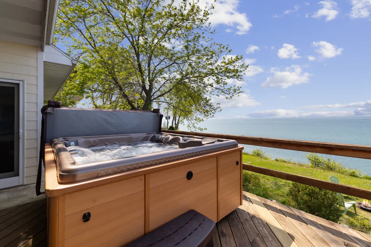 Outdoor hot tub situated on a wooden deck, offering a stunning panoramic view of the expansive lake and lush green landscape under a clear sky.