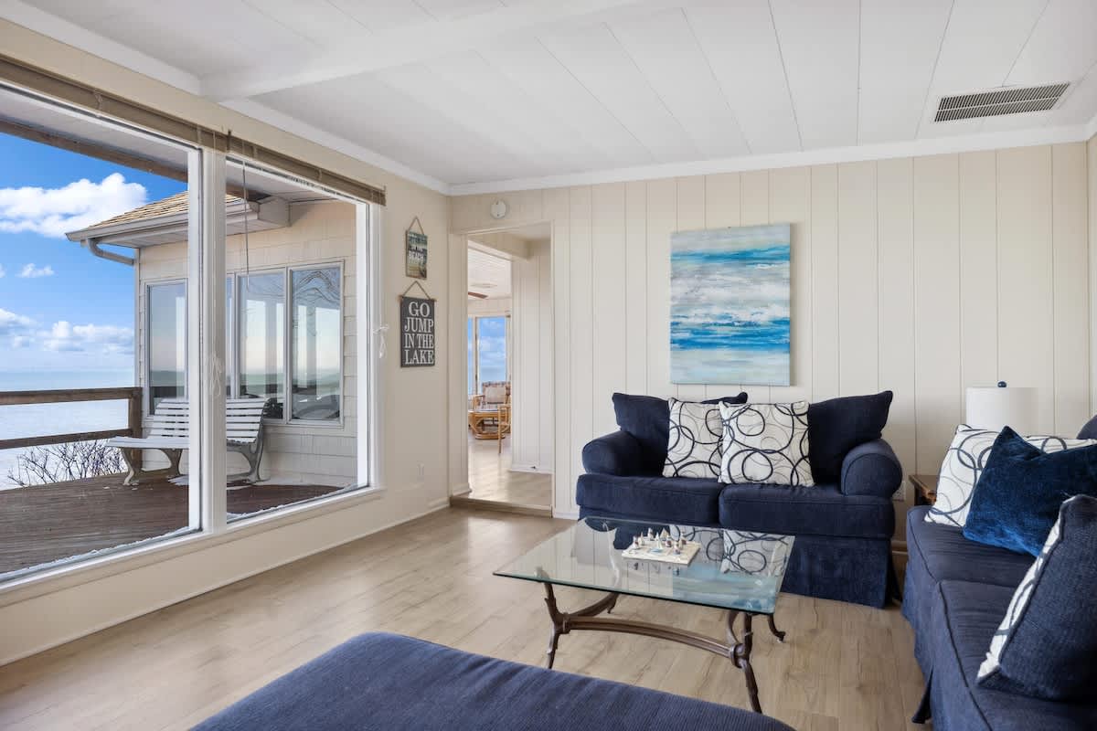 A bright living room with light wood-look flooring showcases a navy blue sofa and a glass coffee table, with large windows offering a clear view into an enclosed sunroom and the serene lake beyond.
