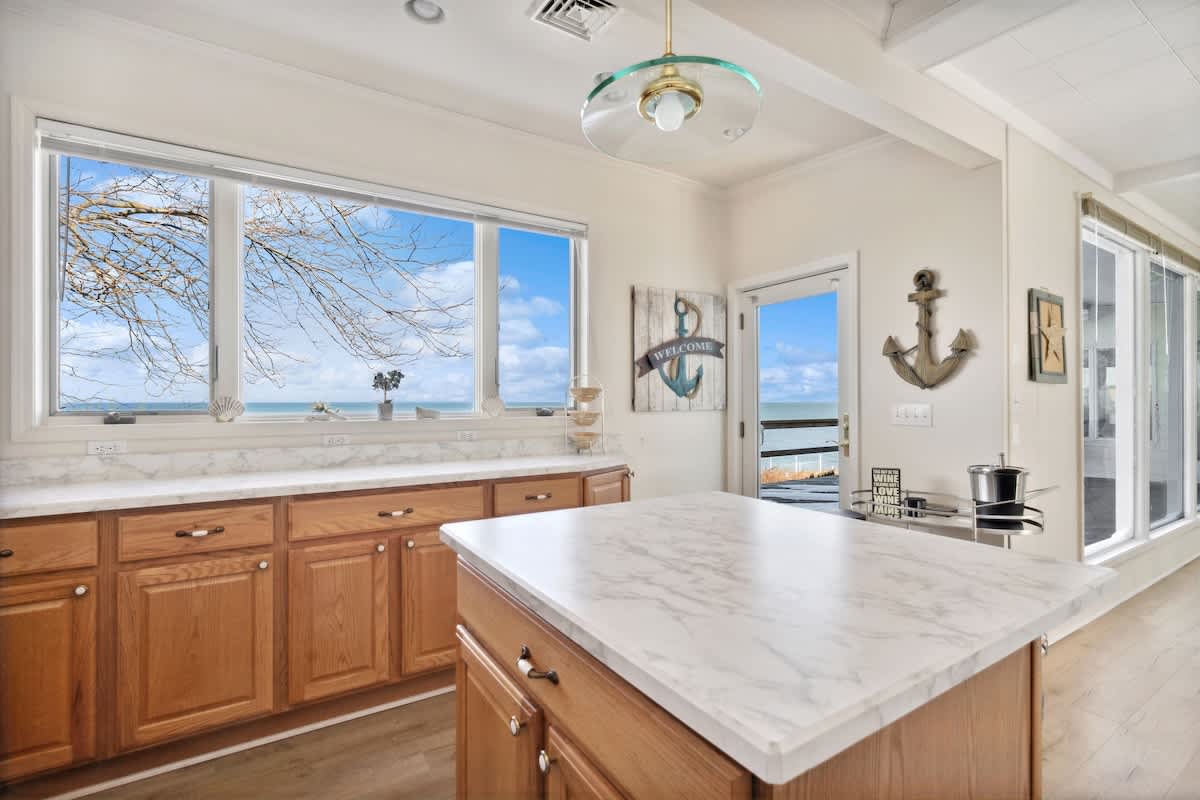A bright kitchen area features light wood cabinets, white marble-look countertops, a central island, and a large window and door offering panoramic views of the lake and direct access to an outdoor deck.
