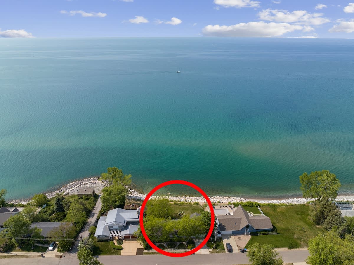Aerial view of a lakefront property, featuring a house with a green yard, situated directly on the shore of a large, blue body of water, with a red circle highlighting the property.