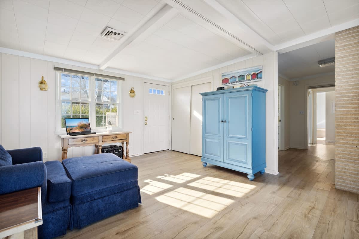 Bright interior space with light wood floors, a blue armoire, a small desk with a laptop, and a blue sofa, receiving ample sunlight through a window.