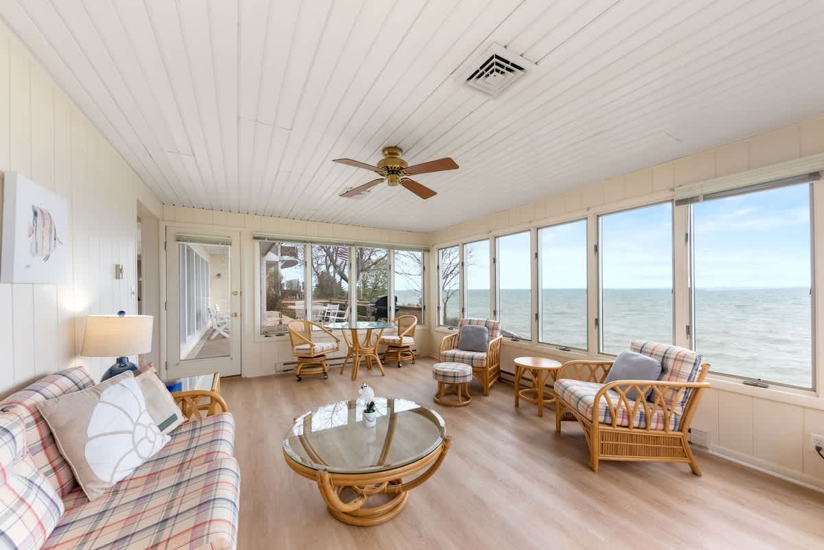A spacious sunroom with light wood-look flooring is furnished with a plaid sofa, wicker armchairs, and a glass coffee table, all set against large windows that provide panoramic views of the lake.