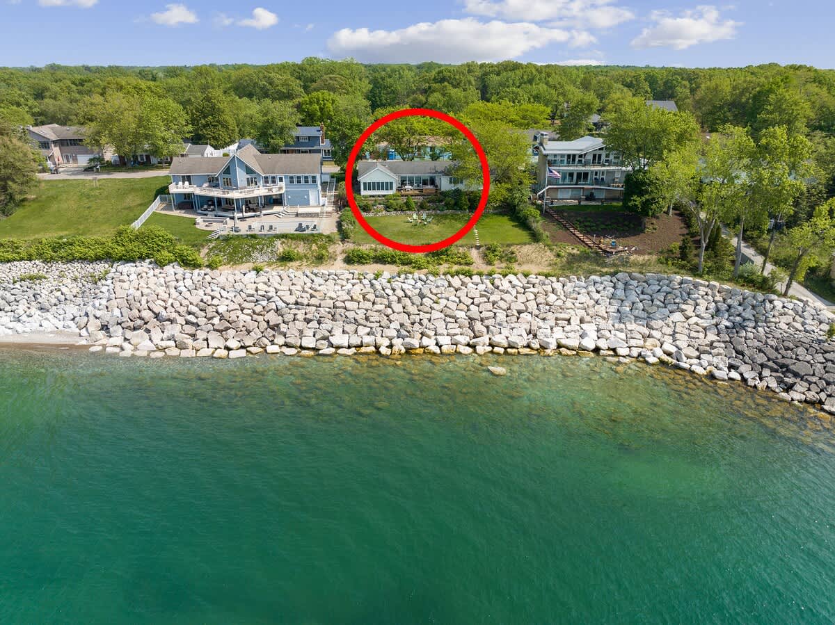 An aerial perspective from the lake highlights the property, circled in red, positioned directly behind a substantial rocky breakwater. The clear turquoise water contrasts with the green landscape and neighboring homes along the shore.
