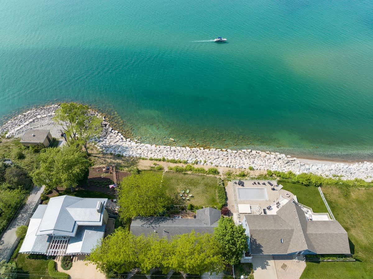 Aerial view showcasing the lakefront property with a house, a well-maintained green lawn, and a boat on the clear blue water of the lake, surrounded by trees.
