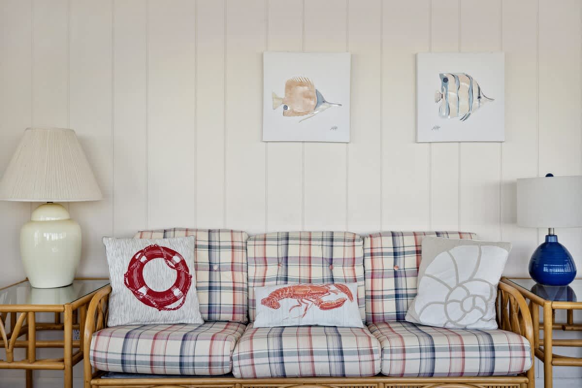 A close-up of a plaid sofa in the sunroom, adorned with nautical-themed pillows (lifebuoy, lobster, seashell), is flanked by end tables with lamps, set against light-colored vertical paneling on the wall.
