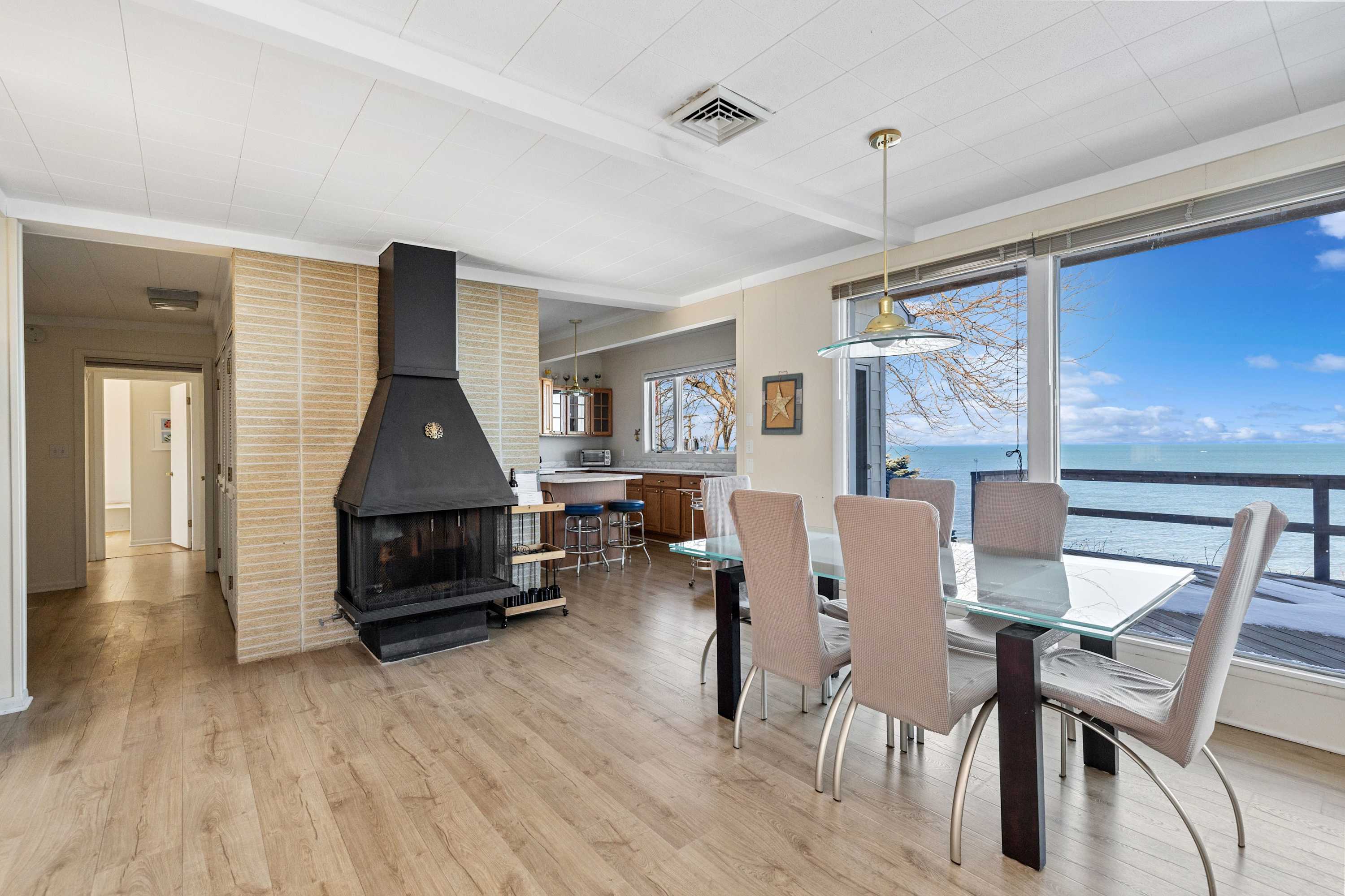 A bright dining area features a glass-top table with four light-colored chairs, set against a large window offering expansive lake views, and a prominent black fireplace with a light-tiled chimney separating it from the kitchen.