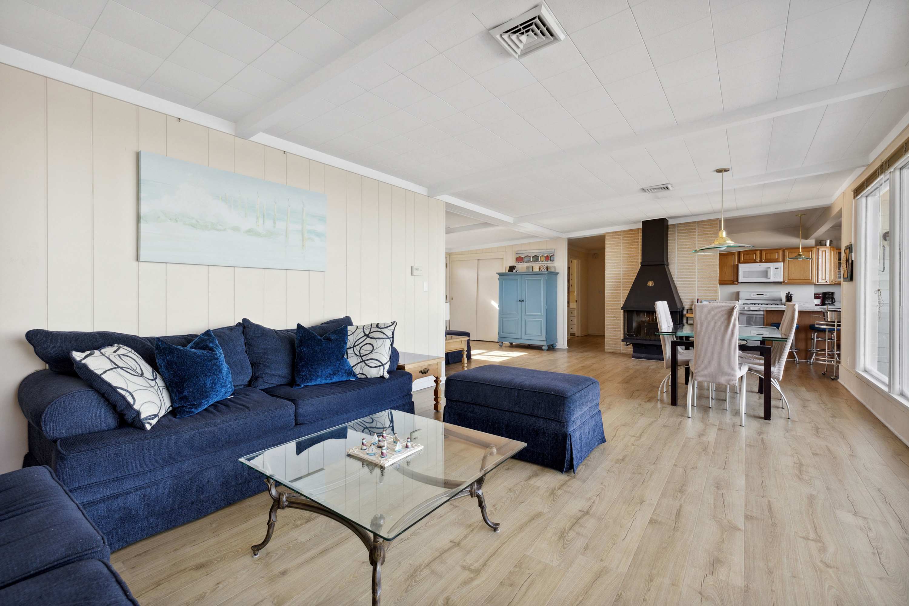 A spacious living room with light wood-look flooring features a large navy blue sofa with decorative pillows and a glass coffee table, offering a view towards the dining area, kitchen, and a large window with a lake view.